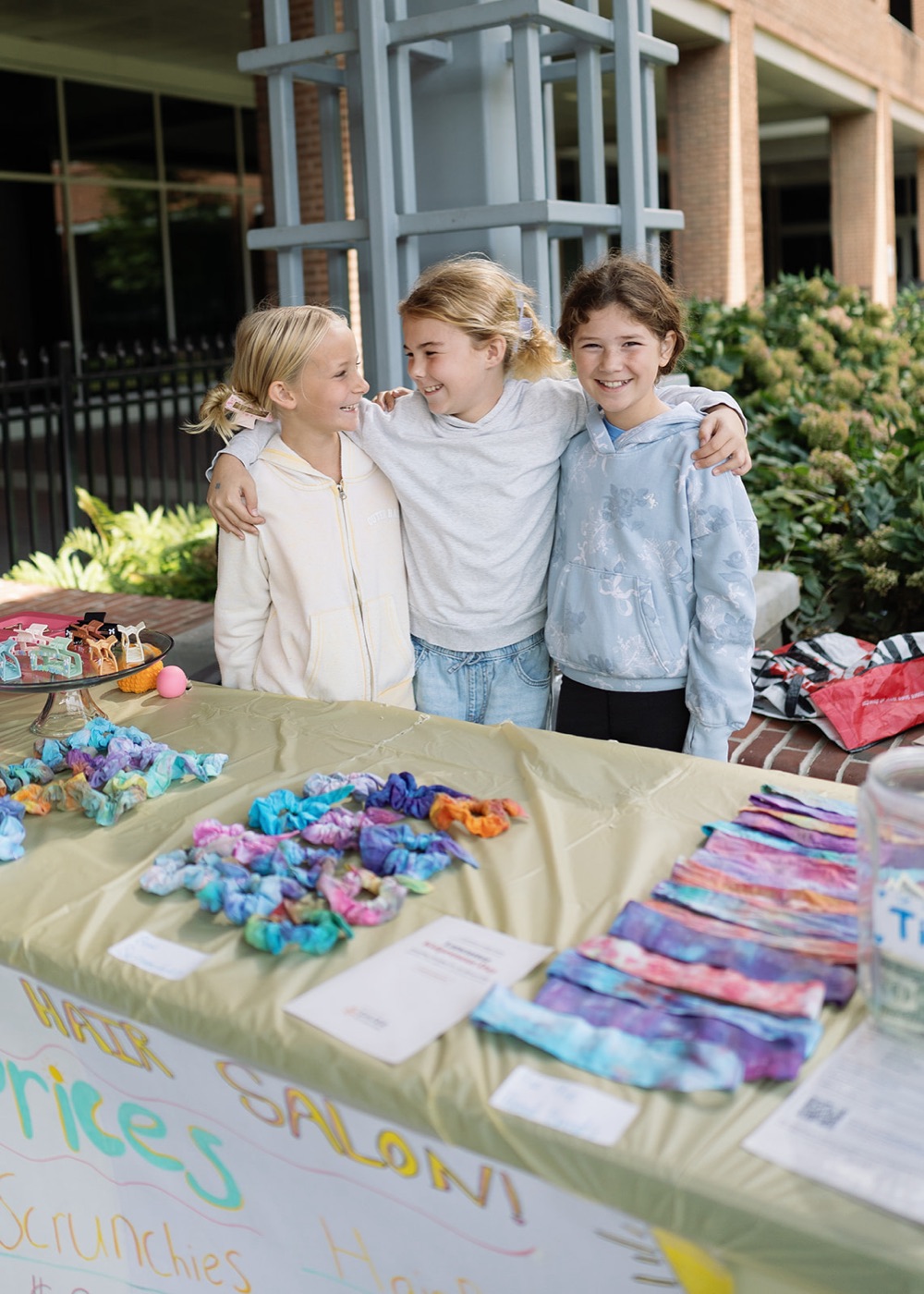 Three kids behind a table of tie-dye scrunchies and hair clips
